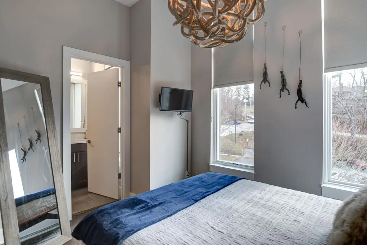 A stylish, inviting bedroom highlighted by a bed adorned with a blue throw and decorative pillows. A unique chandelier and a large mirror elevate the room’s design, while the flat-screen TV and window allow for both entertainment and plenty of natural light, creating a space perfect for both relaxation and privacy.
