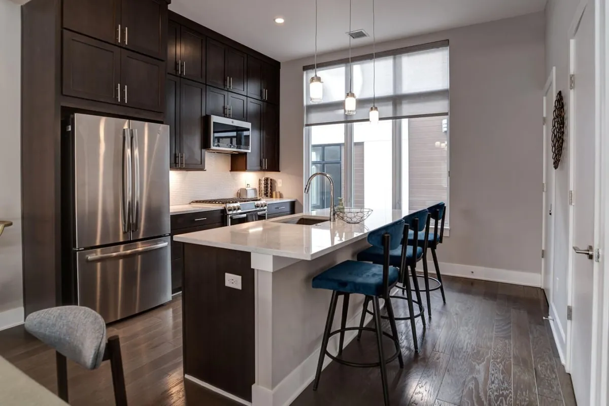 In this image, the kitchen features a large island, stainless steel appliances, bar stools, and expansive windows letting in lots of natural light. The space is open and allows for easy movement, suggesting a layout that is accessible and user-friendly.
