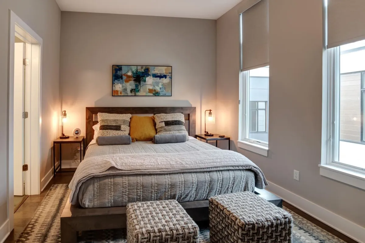 A tranquil bedroom featuring a cozy bed with soft, textured bedding, complemented by tasteful artwork above the headboard and coordinated nightstands with ambient lighting. Sitting stools at the foot of the bed add both style and functionality, while natural light floods the space, enhancing its serene atmosphere.