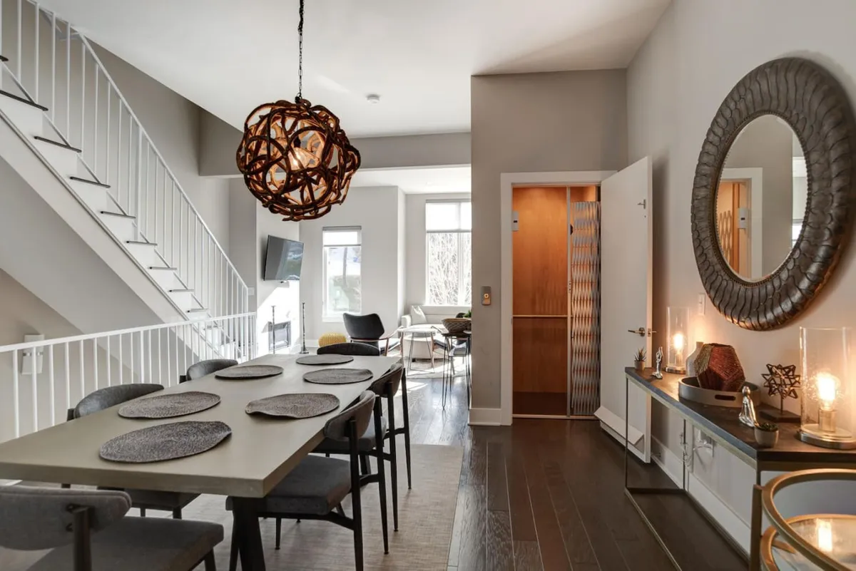 This image features a contemporary, open-plan dining area with a decorative light fixture and mirror, as well as a visible staircase leading to another floor. The space is bright, warm, and welcoming, with clear sightlines that emphasize ease of movement between levels.