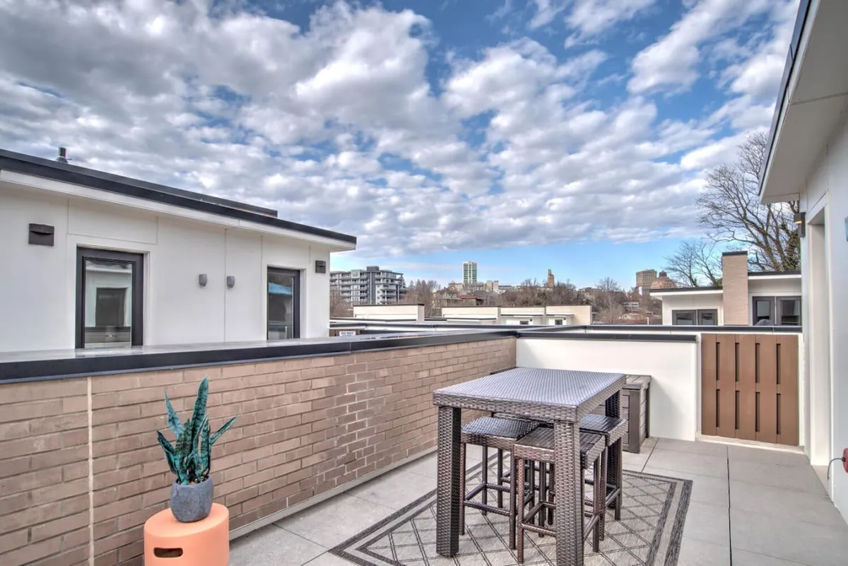 This image features an outdoor dining table set on a rooftop terrace. Potted plants and a cloudy sky add to the ambiance, while city buildings provide a sweeping urban backdrop. The view is clearly elevated, suggesting a private, tranquil space above the city.
