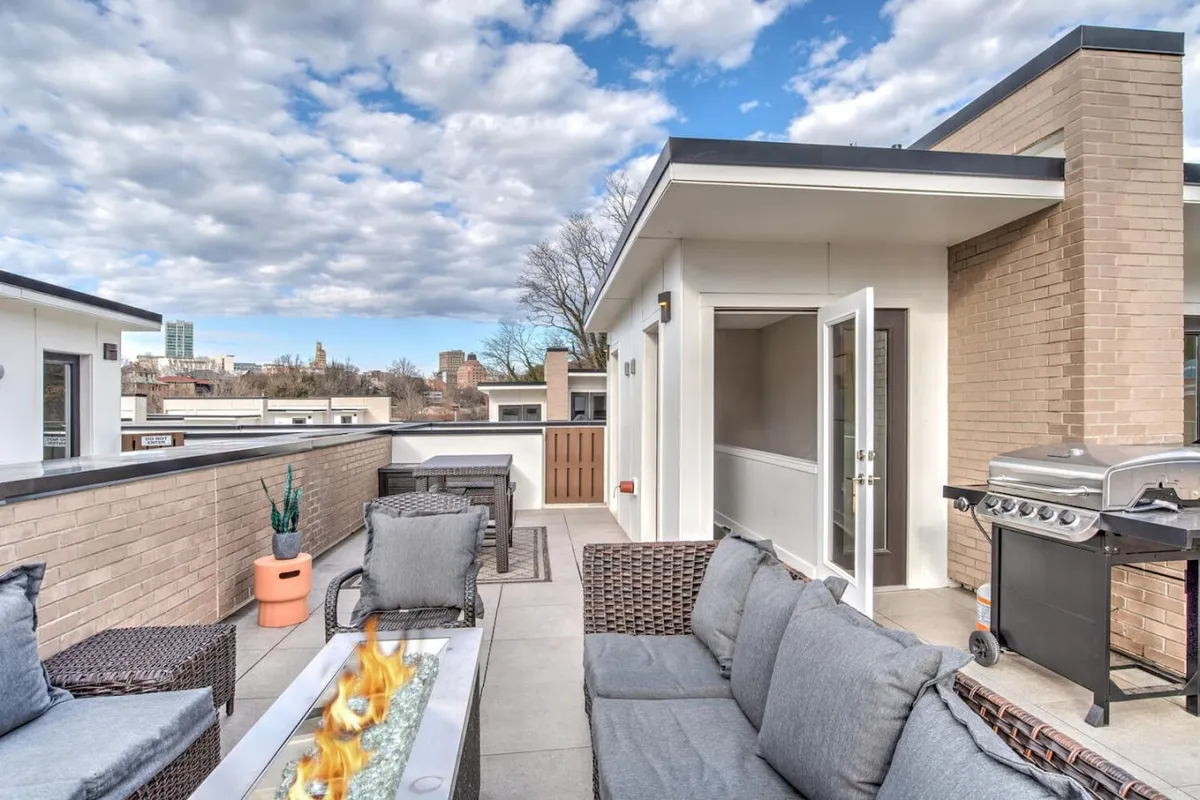 A rooftop scene featuring comfortable outdoor seating, a fire pit, and a grill. The atmosphere is cozy and inviting, ideal for gatherings with friends or relaxing while watching the sunset. The elevation and openness suggest expansive city or mountain views beyond the terrace.