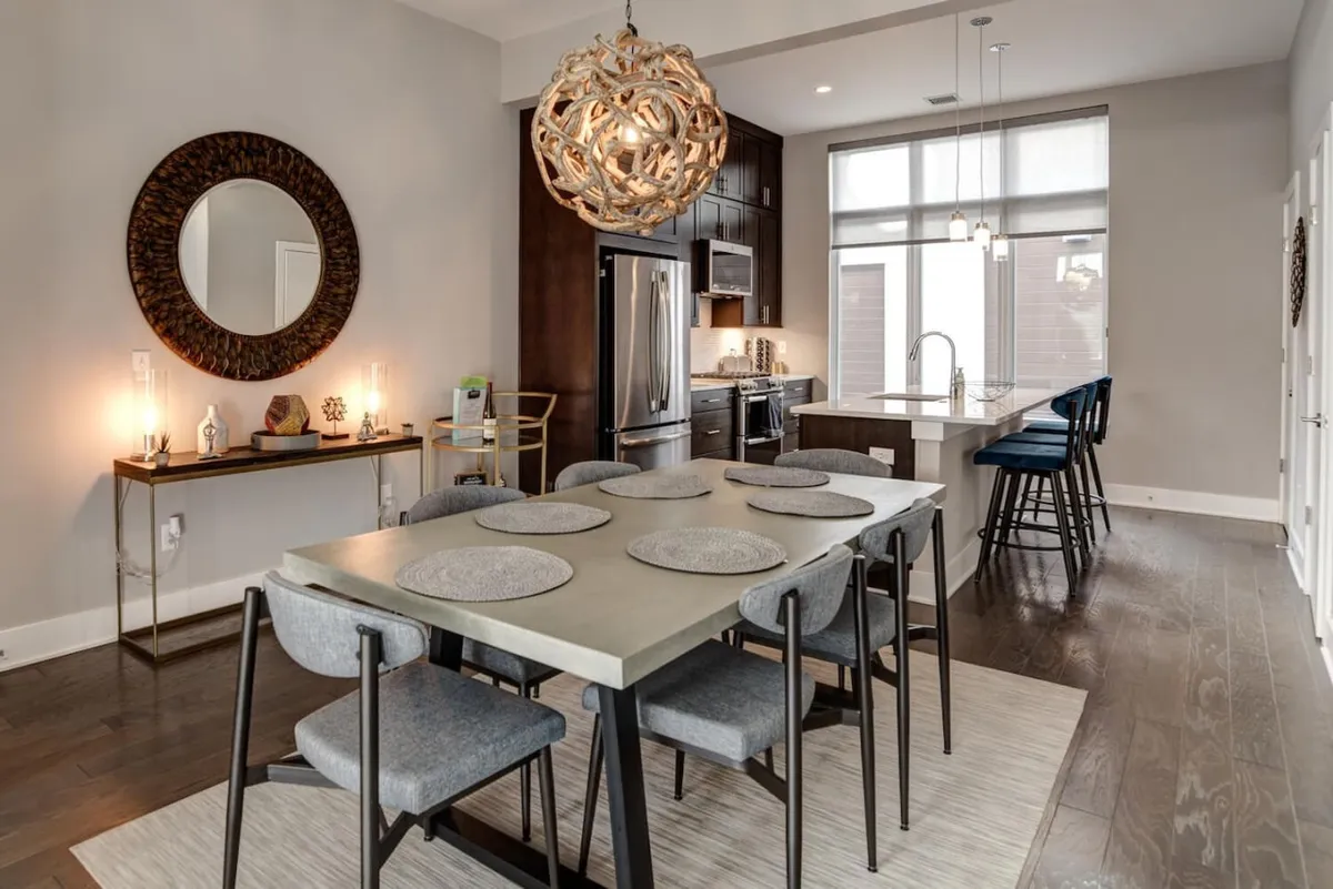 A contemporary dining area with a dining table beneath an artistic chandelier. The space is open, with a view towards the kitchen island and a decorative mirror reflecting light. The setting suggests an inviting environment for shared family meals, entertaining guests, or enjoying quiet mornings.