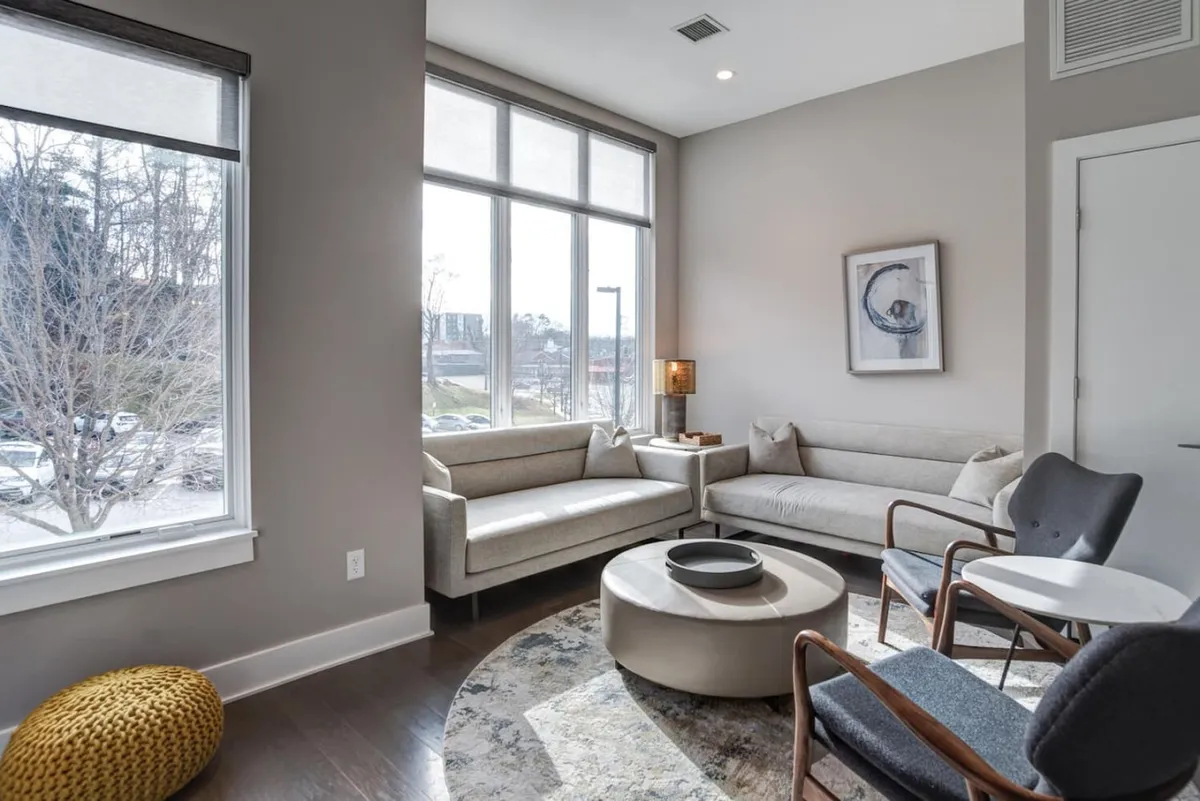 A bright, open living area with large windows flooding the space with natural light. The room features a stylish seating arrangement, a round decorative coffee table, and curated artwork on the walls, creating a comfortable and inviting atmosphere. The sunlight streaming in highlights the serene and welcoming mood of the space, ideal for relaxation or entertaining.