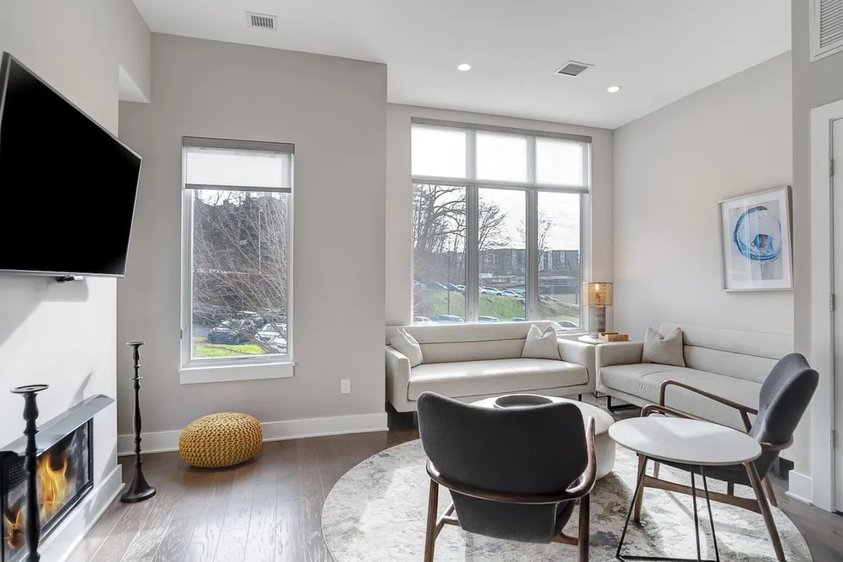 The image showcases the sun-drenched interior of a modern townhome living area featuring expansive floor-to-ceiling windows, contemporary furnishings, a cozy fireplace, and breathtaking mountain and cityscape views beyond. Natural light floods the space, and there's a seamless blend of comfort and style, perfect for relaxing or gathering with friends and family.