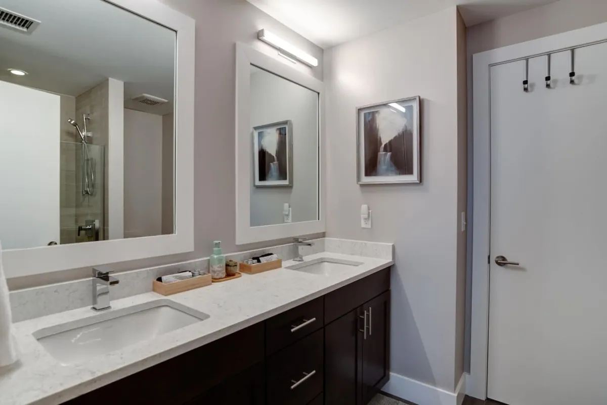 A calming, modern bathroom featuring a double sink vanity, large mirrors that reflect morning sunlight, and elegant wall art. The serene mood is perfect for personal grooming, quiet moments of self-care, or shared routines to start and end each day refreshed.