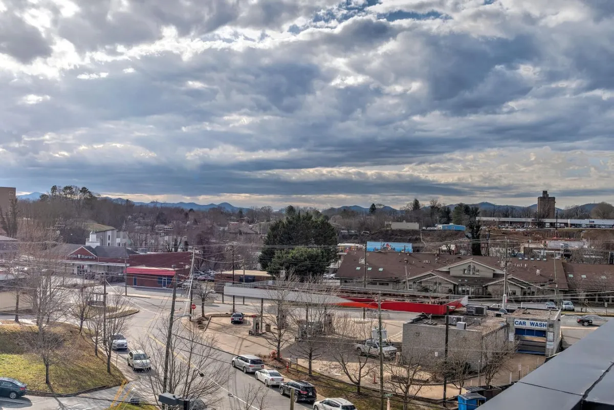 A scenic view of a mountain range in the background, with a street scene featuring commercial buildings, a red building, and local businesses. Trees line the street, and there’s a sense of adventure and exploration, evoking the excitement of discovering Asheville’s Brewery District and its local flavor.